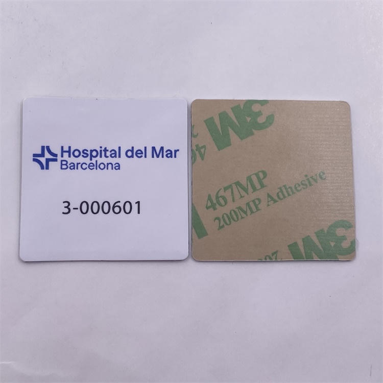 Vista aproximada de uma etiqueta RFID quadrada de PVC impressa para superfícies metálicas ao lado do liner adesivo, mostrando a estrutura de montagem em itens metálicos.